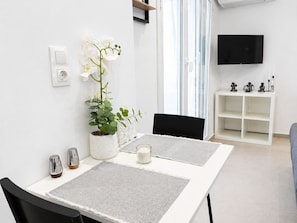 Ein moderner Essbereich mit einem weißen Tisch, zwei Stühlen, einer Topfpflanze, einer Kerze und einem weißen Regal mit Dekoration.