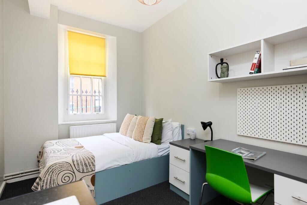 Un dormitorio con una cama, un escritorio, una silla, un radiador, una ventana con persianas y una estantería con objetos.
