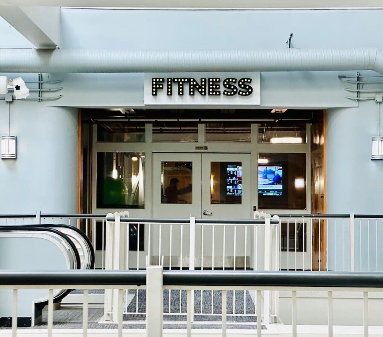 Entrada a un gimnasio con un letrero, puertas de vidrio y una reja de seguridad.
