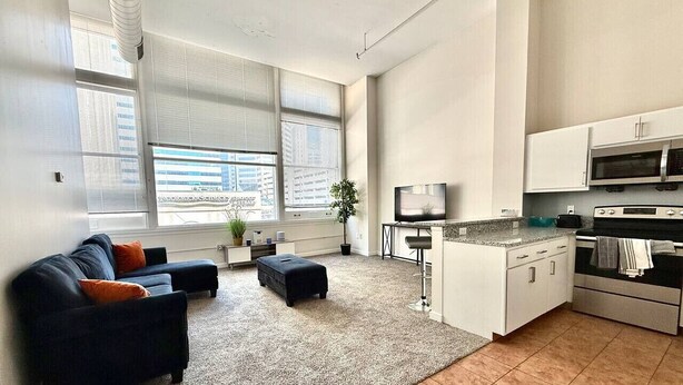 Un salon moderne avec un canapé, un pouf et un téléviseur. Il y a également un coin cuisine équipé d’un four à micro-ondes et d’un four traditionnel.