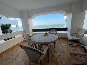 Une table ronde entourée de chaises, sur un balcon donnant sur la mer.