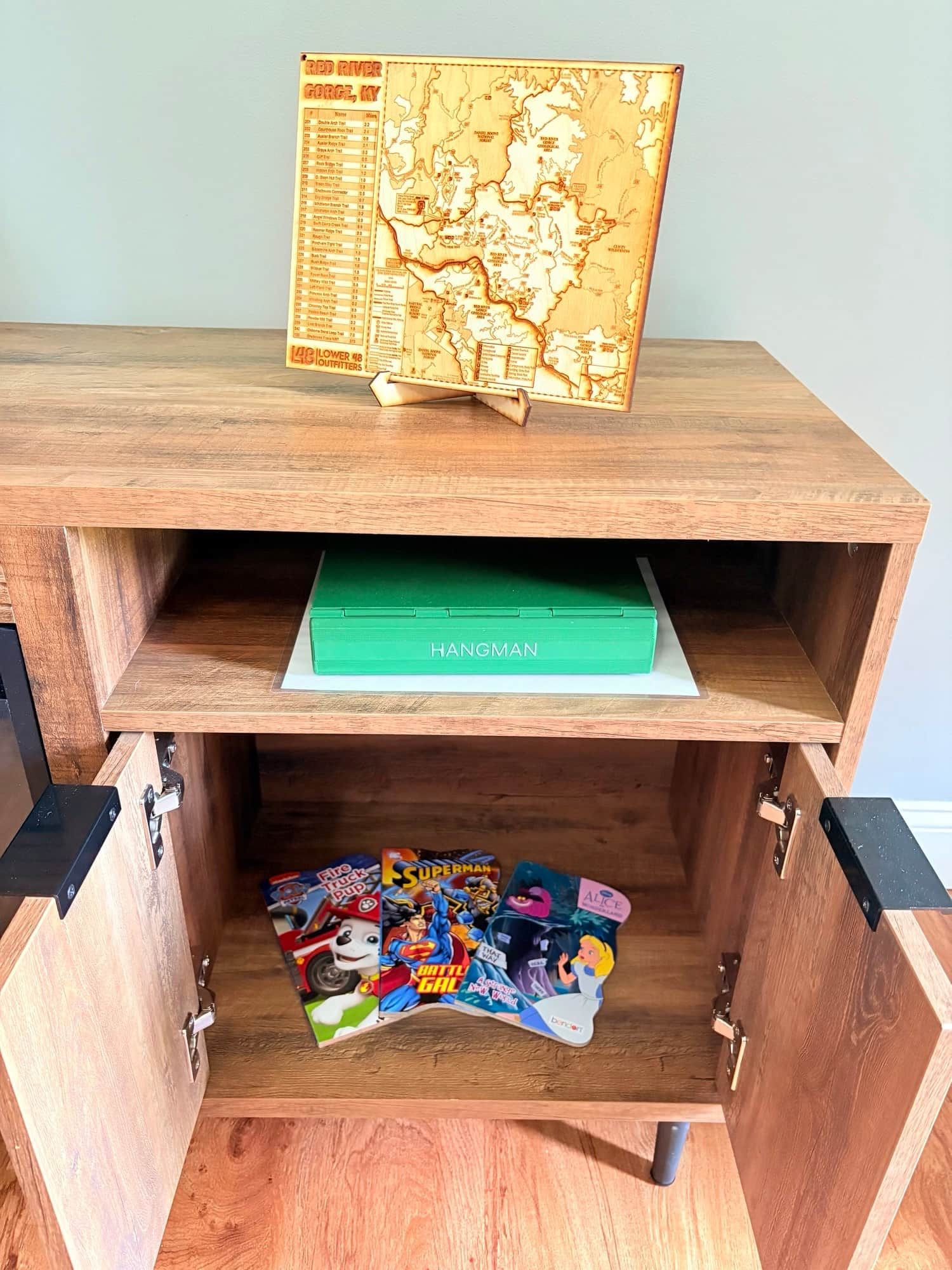 A wooden cabinet with a map on top, a green box labeled 'HANGMAN' in the middle, and a collection of colorful books at the bottom.