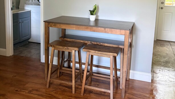 Un coin repas avec une table et un banc en bois, un tableau représentant un paysage et, au fond, une cuisine.