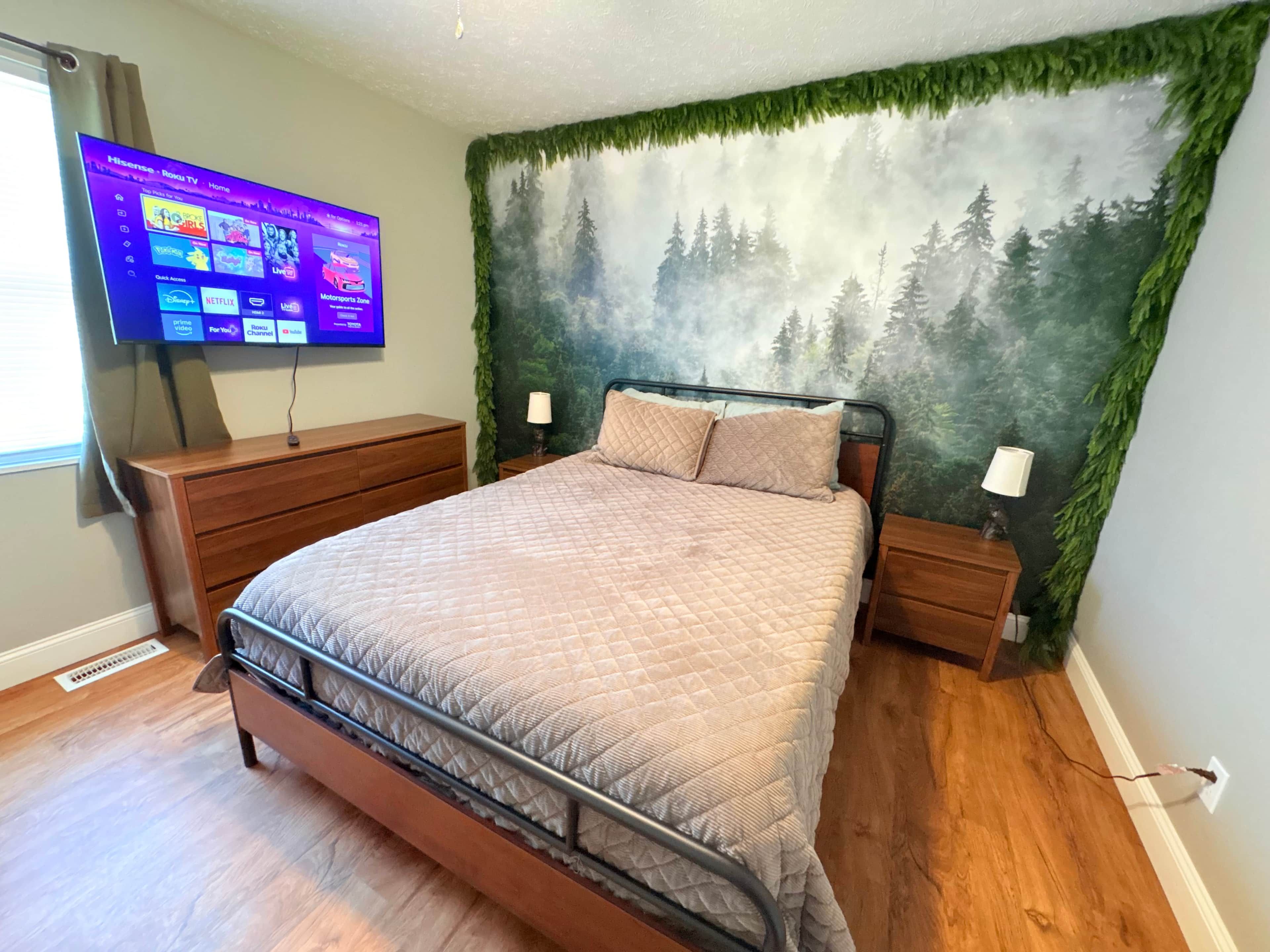 Un dormitorio con cama, cómoda, televisor montado en la pared y un mural con motivos forestales.