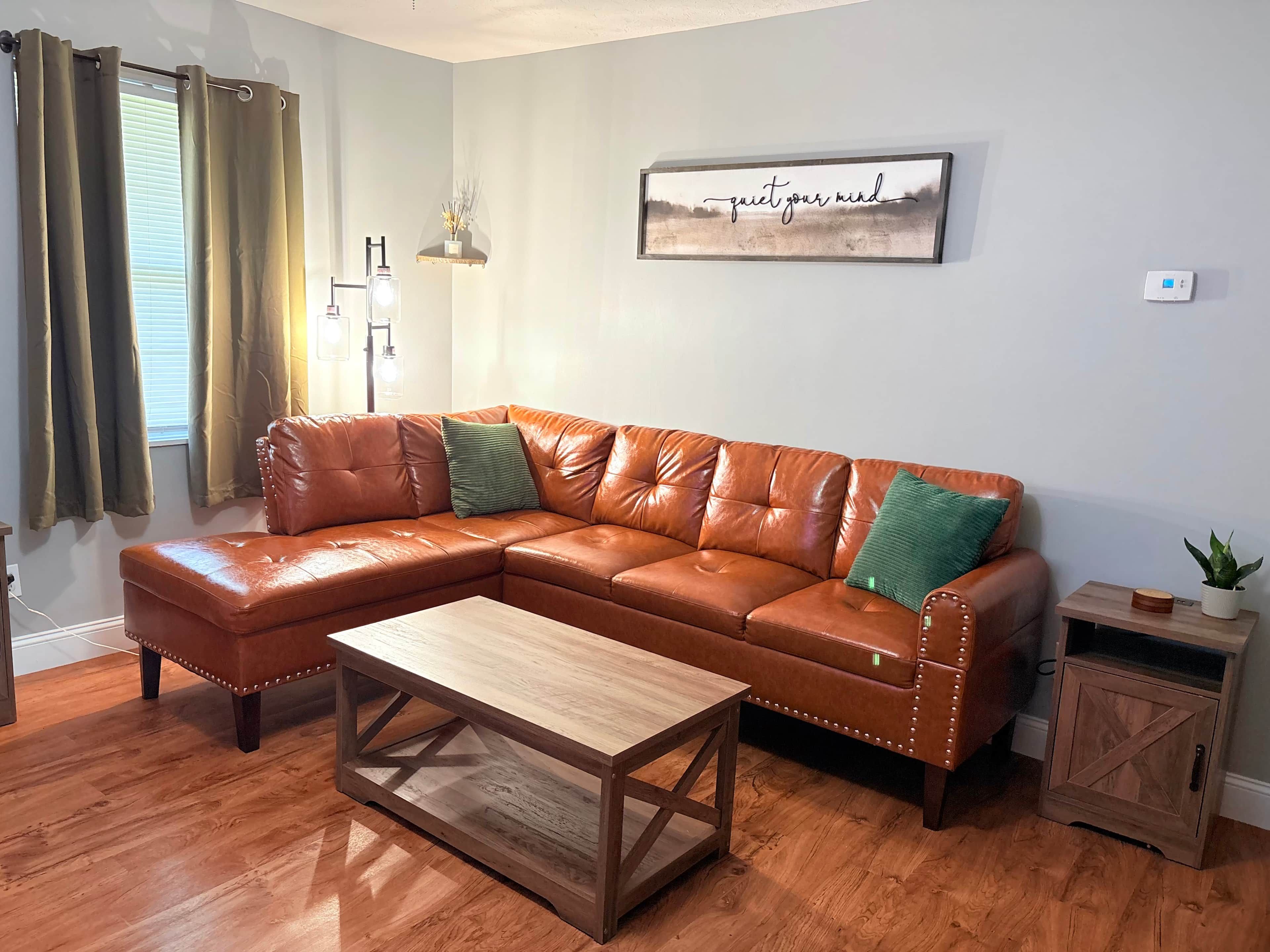 Un salon avec un canapé d’angle en cuir marron, une table basse en bois, un luminaire fixé au mur et une œuvre d’art encadrée portant l’inscription « apaisez votre esprit ».
