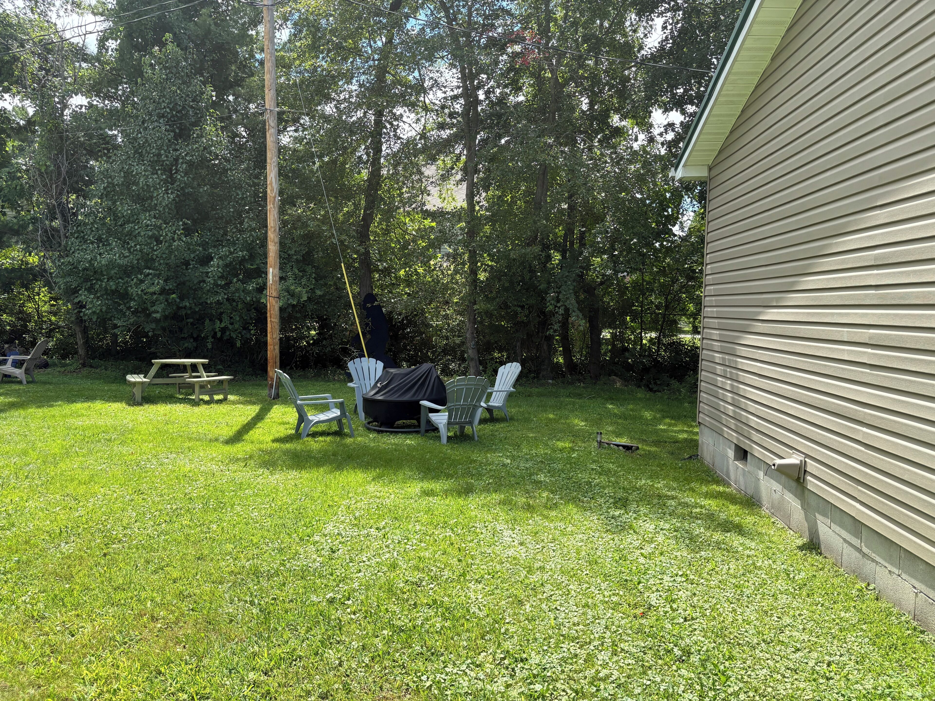 Un patio trasero con una mesa de picnic, sillas Adirondack blancas y una zona de parrilla cubierta.