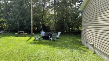 Un patio trasero con una mesa de picnic, sillas Adirondack blancas y una zona de parrilla cubierta.