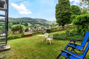 Un espacio al aire libre con césped, una mesa y sillas, un sillón azul y vistas a un pueblo y colinas al fondo.