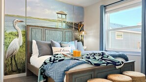 Un dormitorio con cama, almohadas, una mesita de noche con un vaso de jugo de naranja, una lámpara y una ventana con cortinas.