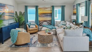 Una sala de estar con un televisor grande, un ventilador de techo, un conjunto de sofás y vista al océano a través de las ventanas.