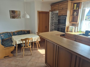 Une cuisine avec des meubles en bois, une table à manger avec des chaises, un four encastré et une fenêtre avec des rideaux.