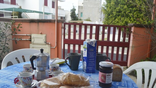 Uma mesa com toalha azul, um coador de café, um leite, um pote de geleia e um prato de pão.
