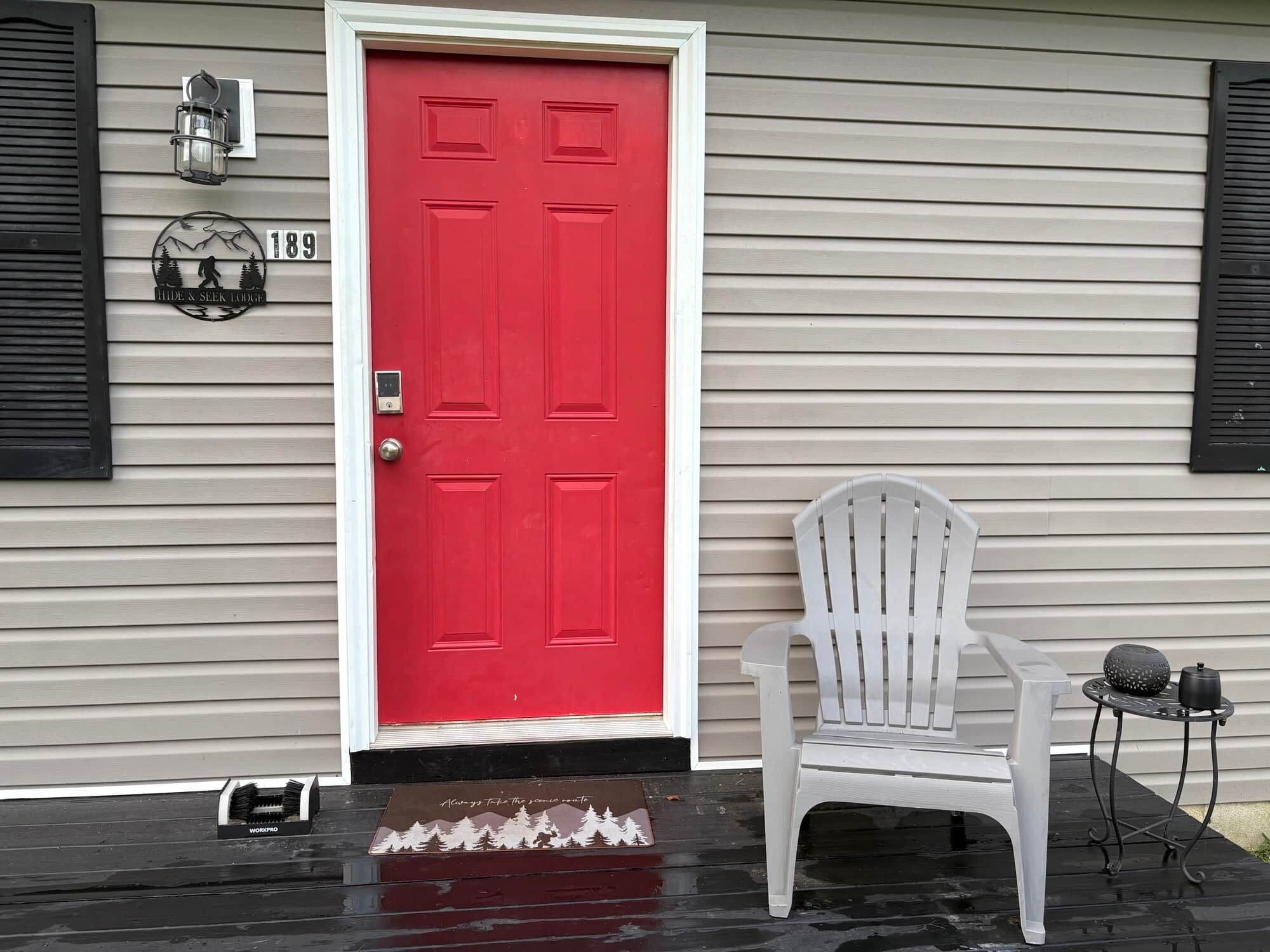 A red door with a mailbox and a white chair outside a house with siding.