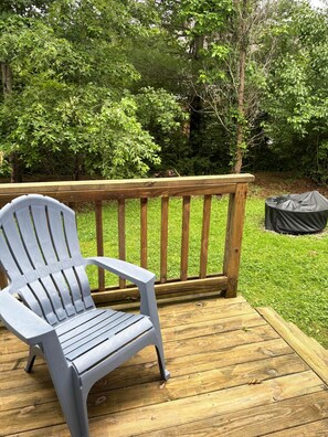 A wooden deck with a grey Adirondack chair, surrounded by lush greenery and trees.