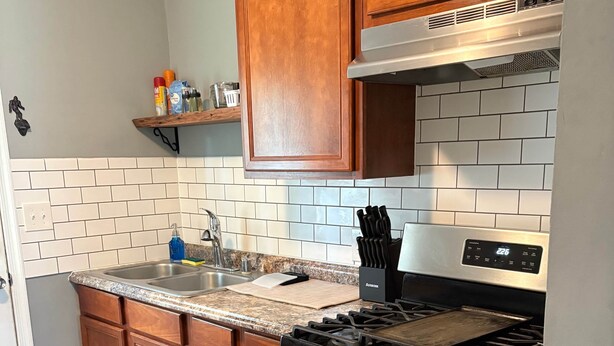 Et køkken med træskabe, komfur i rustfrit stål, vask og flisebeklædt backsplash.