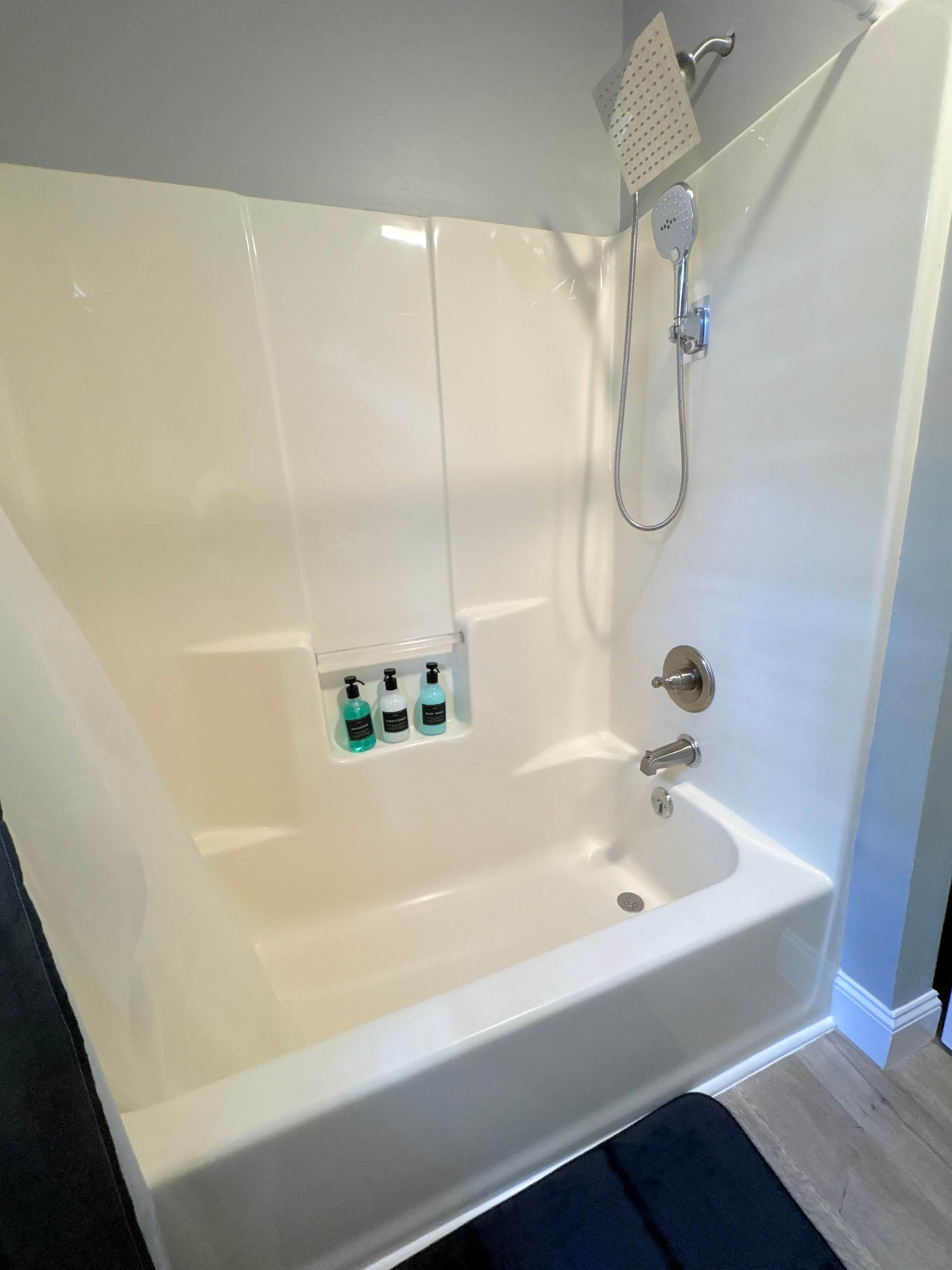 A white bathtub with a handheld showerhead and a built-in faucet. There are three bottles of liquid soap in a shelf below the tub.