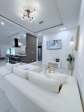 Ein modernes Wohnzimmer mit einem weißen Sofa, einem Essbereich, einer Küche und einem an der Wand montierten Fernseher.