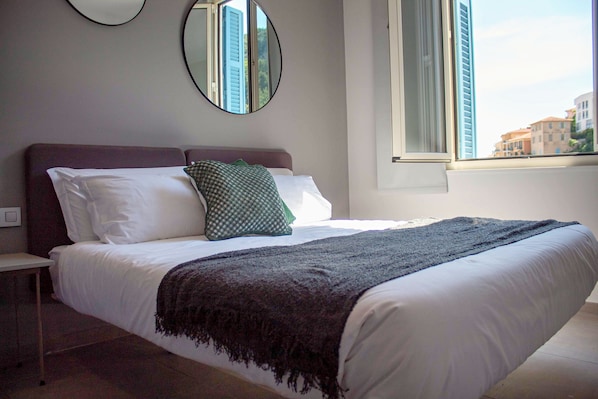 A bedroom with a bed, a nightstand, a window with a view, and a mirror on the wall. - Appartement 6B - MyResidenced'Azur (Beausoleil)