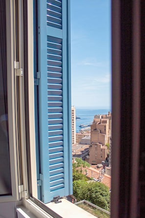 A view through a window with blue shutters, overlooking a coastal cityscape with buildings and the sea. - Appartement 6B - MyResidenced'Azur (Beausoleil)