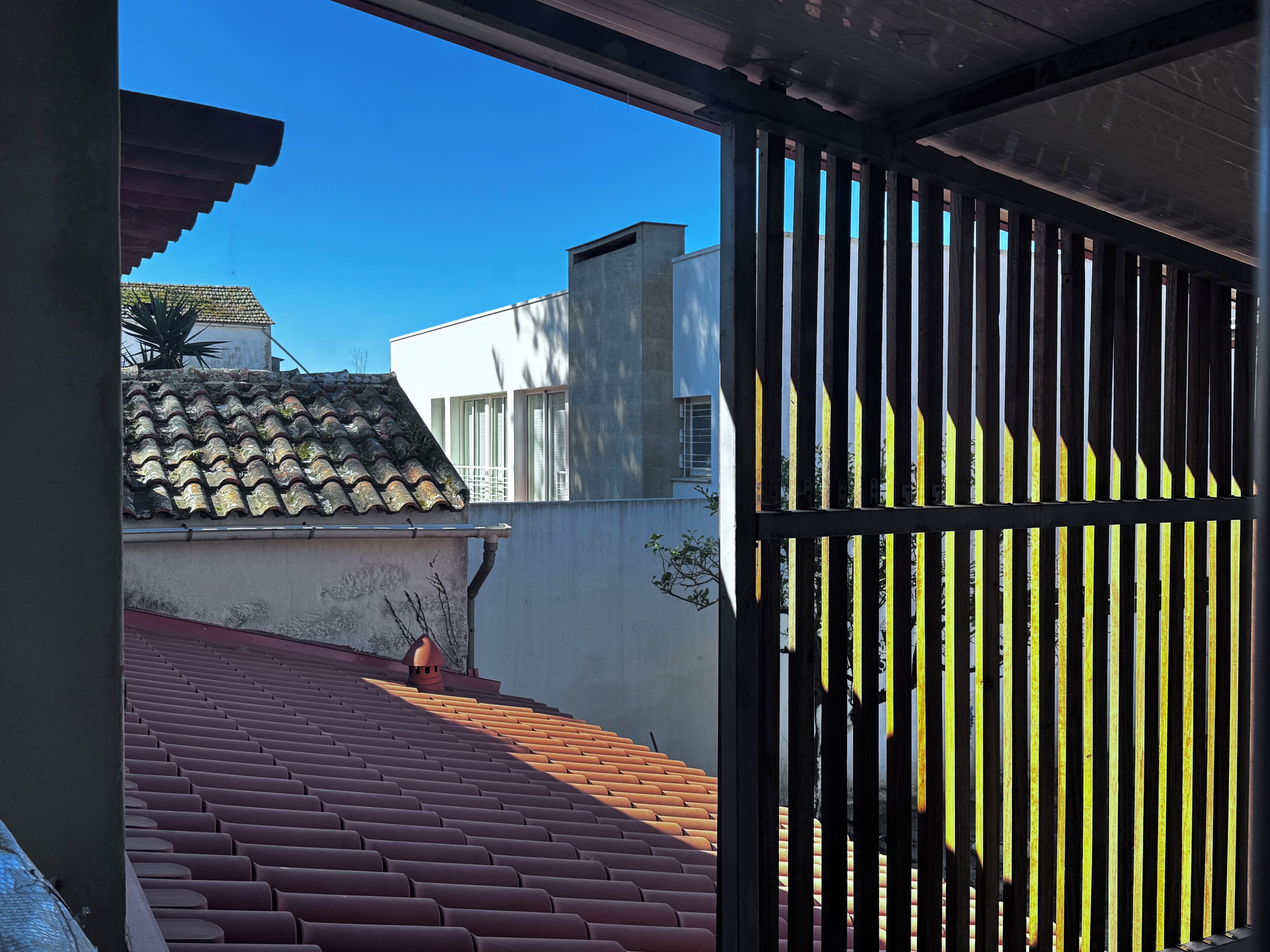 Visão através de um portão de metal, mostrando um telhado de telhas vermelhas, um edifício branco e um céu azul límpido.