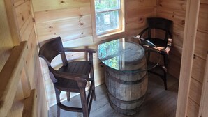 Interior d'una cabana de fusta amb una taula rodona, dues cadires, un barril de fusta i una finestra.
