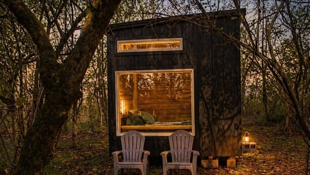 Una petita cabana de fusta amb dues cadires orientades cap a una finestra gran, envoltada d'arbres.