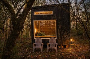 Una cabaña de madera pequeña con dos sillas mirando a un ventanal grande, rodeada de árboles.