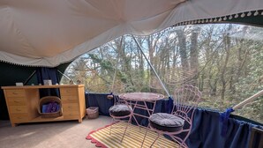 Una habitación con carpa, un tocador de madera, una silla metálica con cojines, una alfombra a rayas y vista a los árboles.
