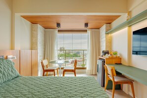 A bedroom with a bed, a desk, a chair, a table, and a view of the city through large windows. - RV12 Beautiful and Cozy Studio in Rio Vermelho (Salvador)