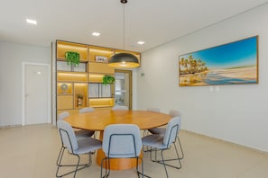 A modern dining area with a round table, six chairs, a pendant light, a wall-mounted painting, and built-in shelving with plants. - RV12 Beautiful and Cozy Studio in Rio Vermelho (Salvador)