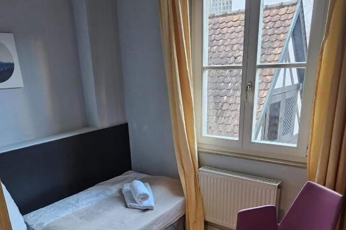 A small, tidy room with a bed, a chair, a radiator, and a window with curtains.