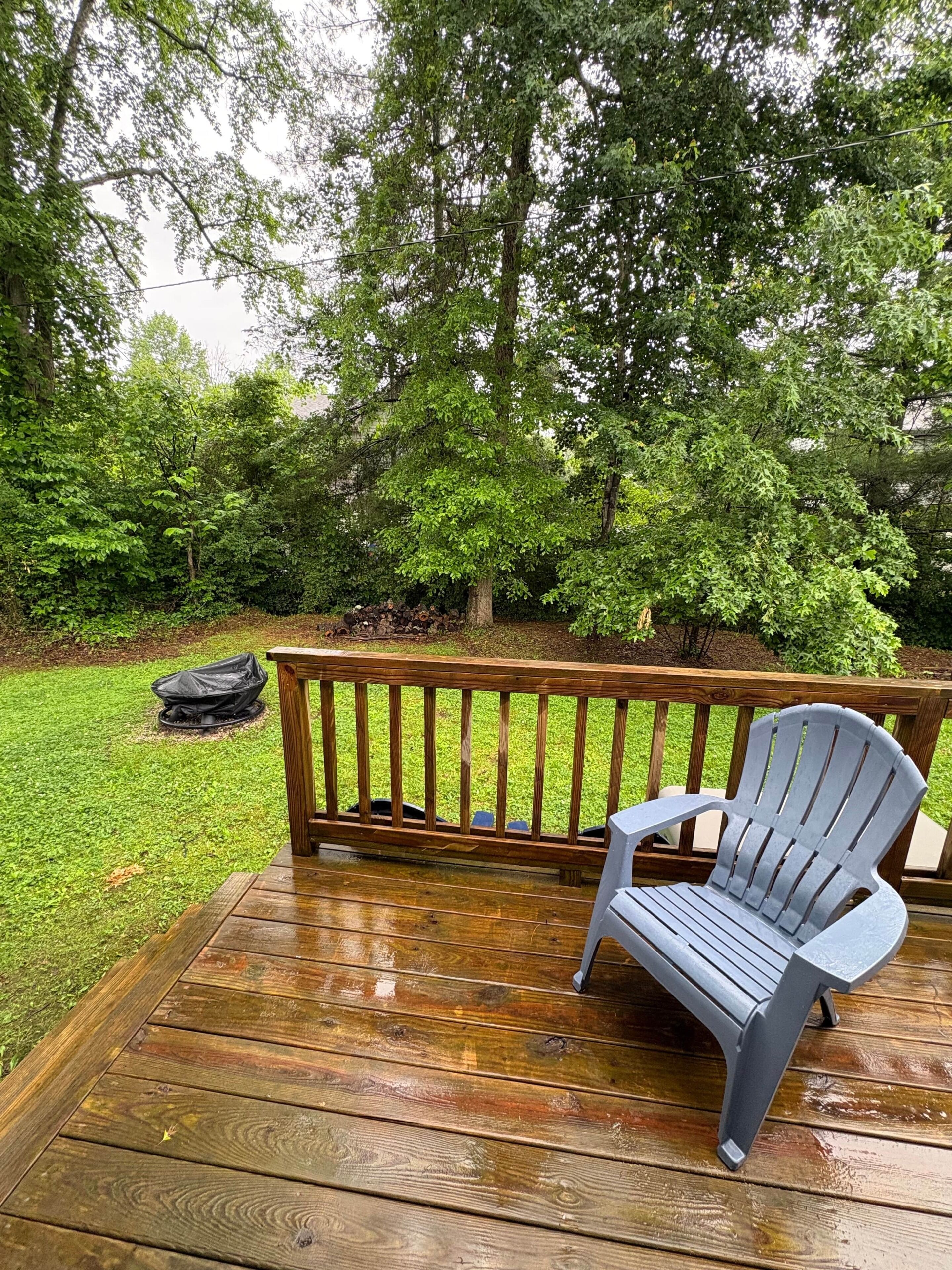 En träveranda med en grå Adirondack-stol, ett träräcke och en hög med ved.