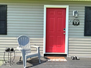 A red door with a mailbox and a number plate, a white chair, and a small table with a kettle and a pot.