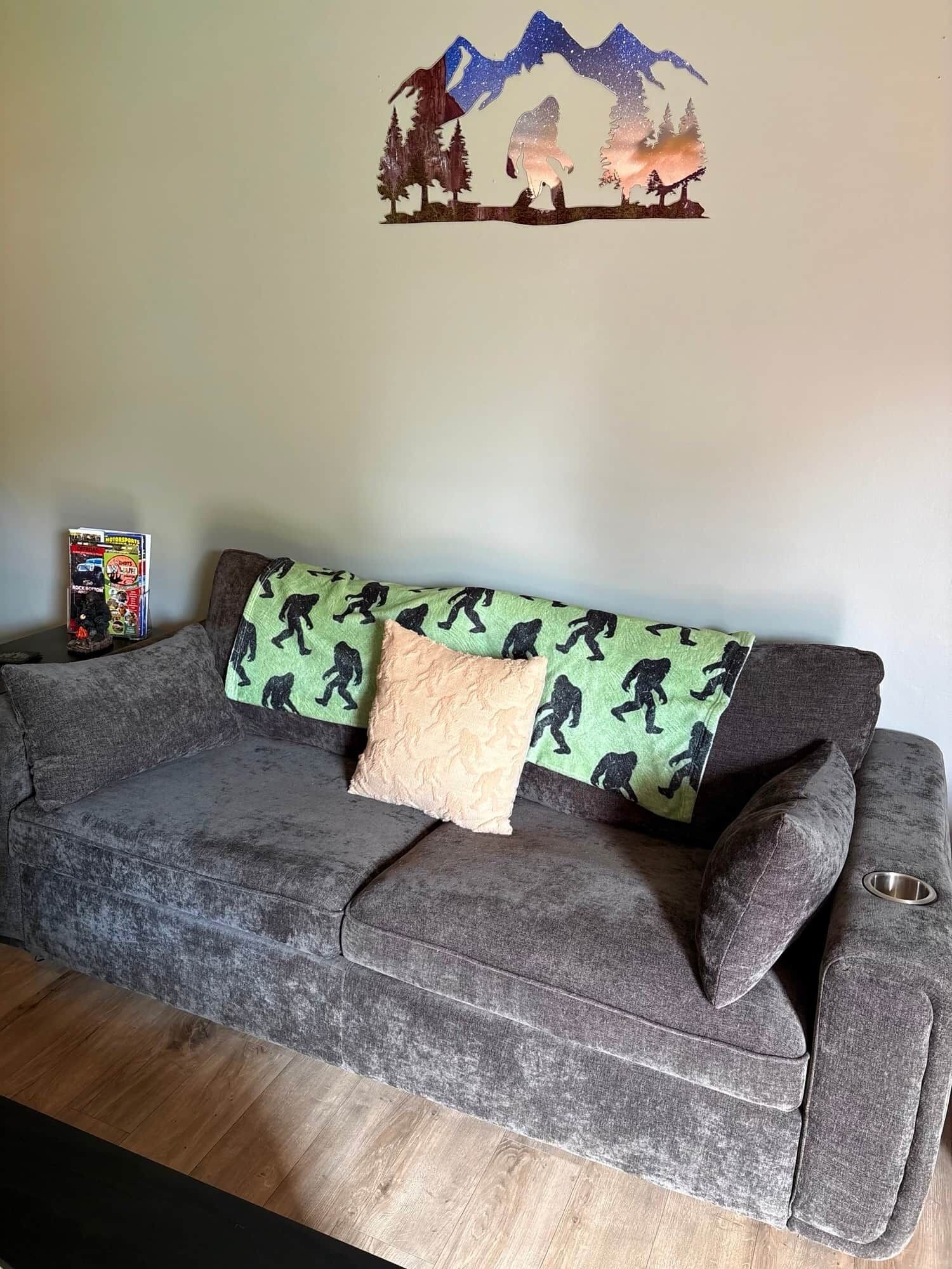 A grey sofa with a green blanket featuring black dinosaur prints, a beige pillow, and a wall decoration of a mountain scene.
