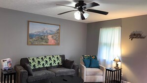 Een woonkamer met een grijze bank, een beige fauteuil, een salontafel, een plafondventilator en een schilderij aan de muur.