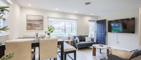 A modern living room with a dining area, a sofa, a TV mounted on the wall, and a window providing natural light.
