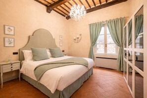 Ein Schlafzimmer mit Bett, Nachttischen, einer Chandelierleuchtre, einem Fenster mit Vorhängen und einer Wanduhr.
