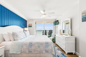 A bedroom with a bed, a dresser, a ceiling fan, a TV, and a balcony with chairs. - Sand Castle Oceanfront Retreat (Indian Shores)