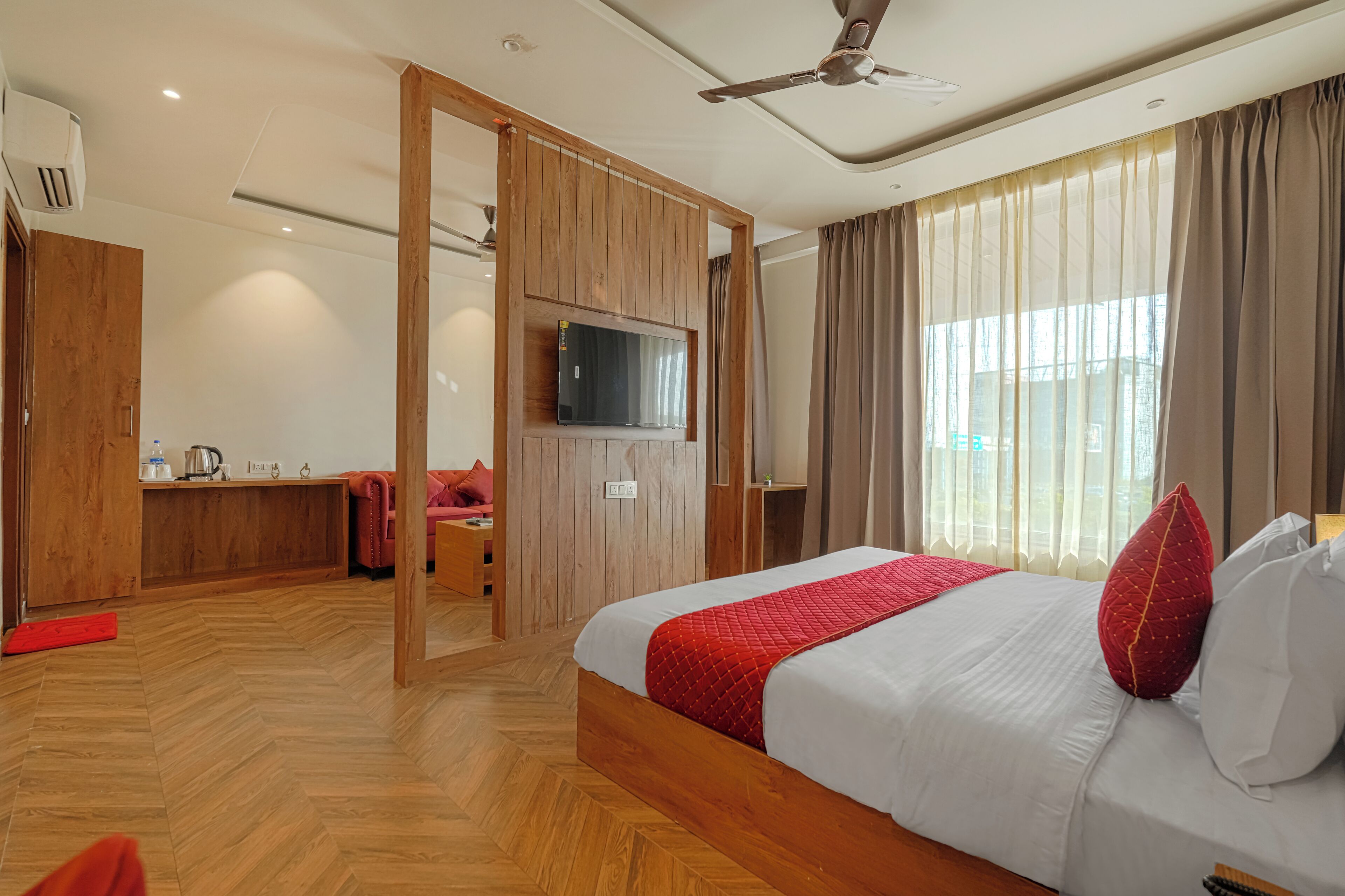 Habitación de hotel con piso de madera, cama con manta roja, televisor montado en la pared y ventana con cortinas.