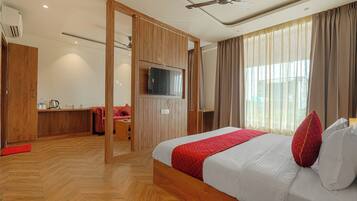 Habitación de hotel con piso de madera, cama con manta roja, televisor montado en la pared y ventana con cortinas.