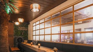 A bar with wooden benches and a long counter, large windows, and hanging pendant lights. - Hotel Escala - Near Golden Gate Amritsar (Amritsar)