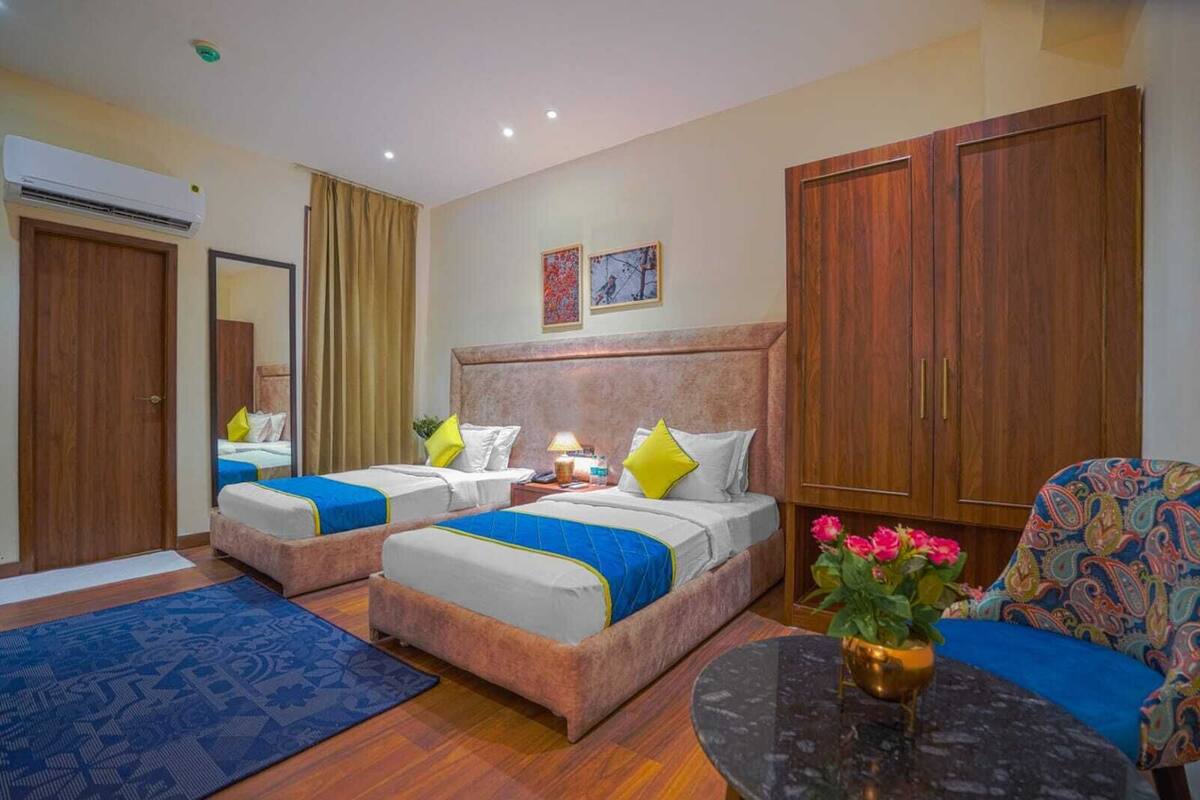 Habitación de hotel con dos camas, una zona de estar, un espejo, un armario y una pequeña mesa con un jarrón de flores.