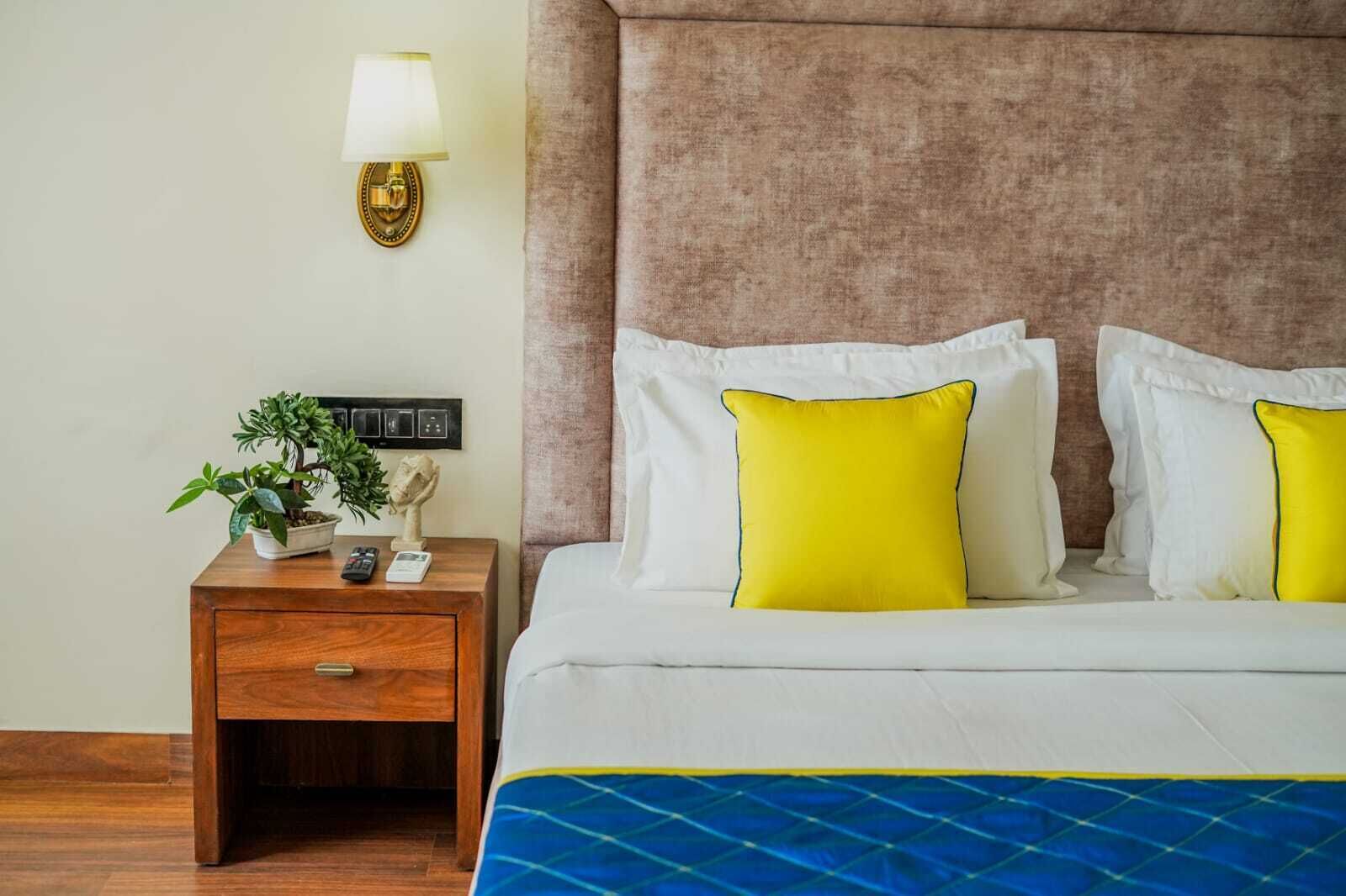 Una cama bien hecha con almohadas blancas y amarillas, una mesita de noche de madera con una lámpara y una planta en maceta.