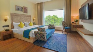 A hotel room with a large bed, bedside tables, a desk, a chair, a TV, and a window with a view of trees. - Hotel Escala - Near Golden Gate Amritsar (Amritsar)