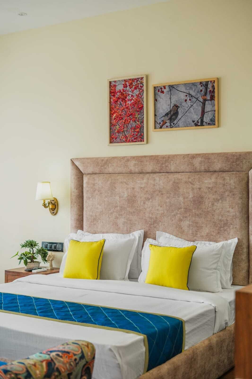 Ein Hotelzimmer mit einem großen Bett, zwei an der Wand befestigten Kunstwerken, einer Nachttischlampe, einem Nachttisch mit Pflanze und einer gemusterten Tagesdecke.