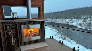 Eine Holzterrasse mit Kamin, Glasbrüstung und Blick auf einen Fluss und eine verschneite Landschaft.