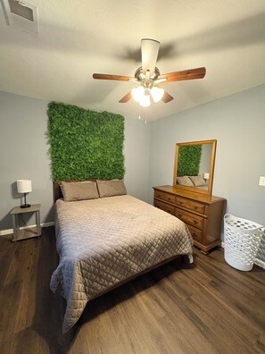 A bedroom with a bed, a dresser, a mirror, a ceiling fan, a wall-mounted green plant, a nightstand, and a laundry basket.