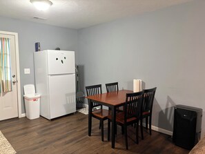 A compact kitchen with a white refrigerator, a trash bin, a dining table with chairs, and a small black speaker.