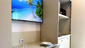 A modern kitchen with a flat-screen TV displaying a beach scene, a dining table with plates and cutlery, and a white chair. - Appartement 4A - MyResidenced'Azur (Beausoleil)