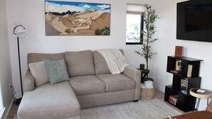 Una sala de estar con sofá, un cuadro, una planta, una estantería y un televisor.